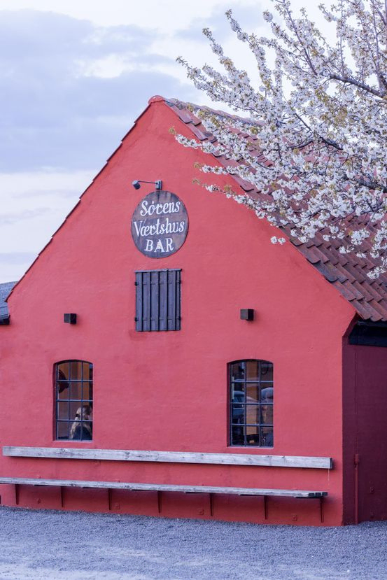 Sørens Værtshus i Snogebæk på Bornholm, hvor man kan spise lækker pizza, høre live-musik og slå søm-konkurrence. Den røde facade ses med Sørens Værtshus skilt og et blomstrende kirsebærtræ står ved siden af.