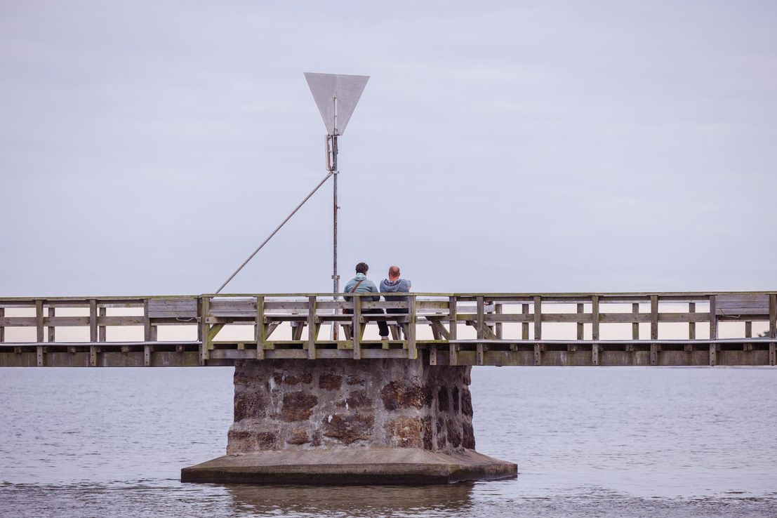 Par sidder på bænk på broen som fører til Snogebæk Havn og nyder udsigten.